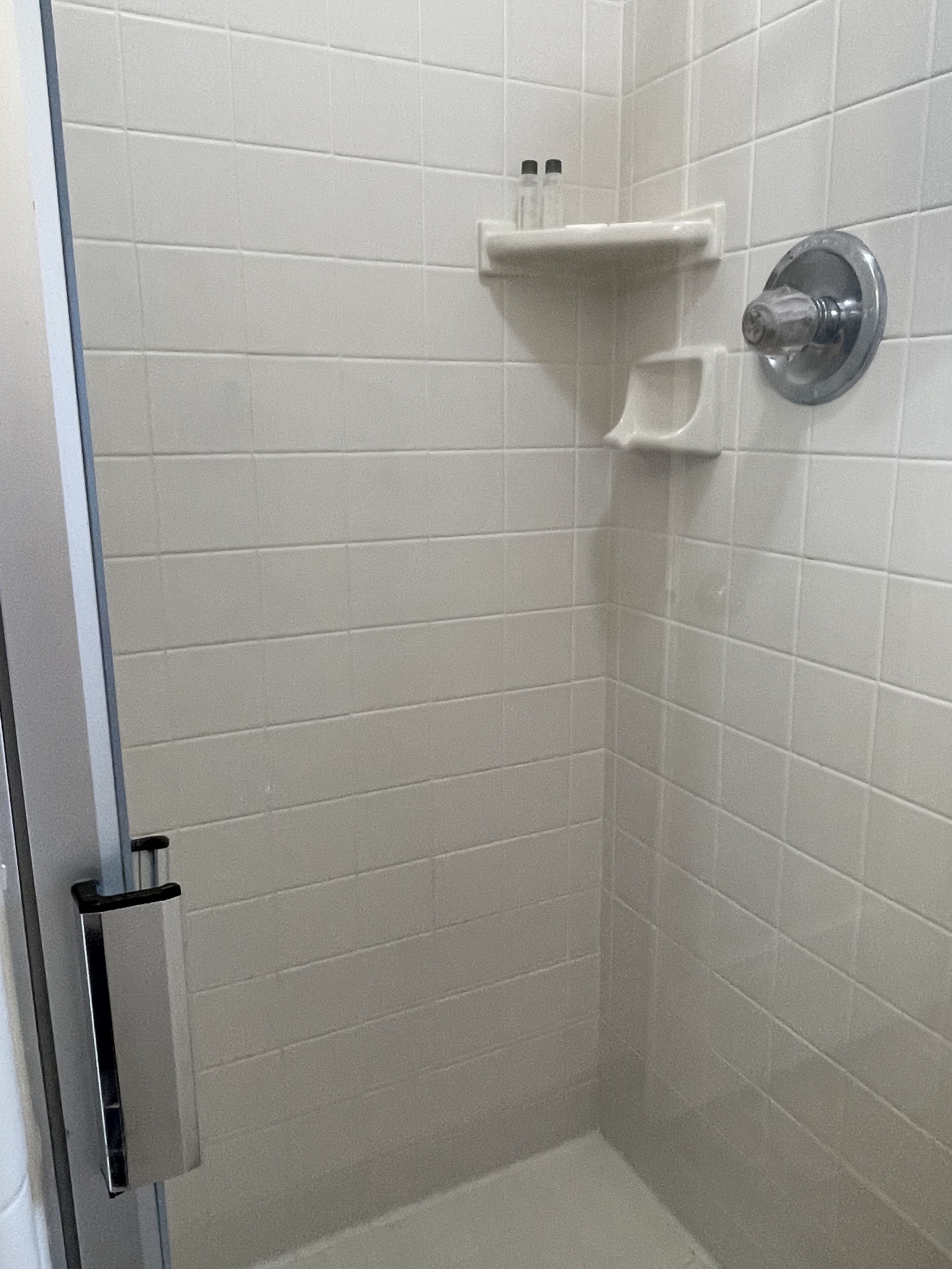 A bathroom with white tiled walls, a shower area, and a shelf with toiletries.