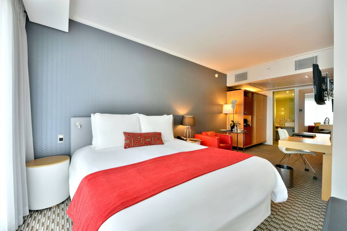 Une chambre d’hôtel avec un grand lit, un bureau, une chaise et une salle de bain visible à travers une porte ouverte.