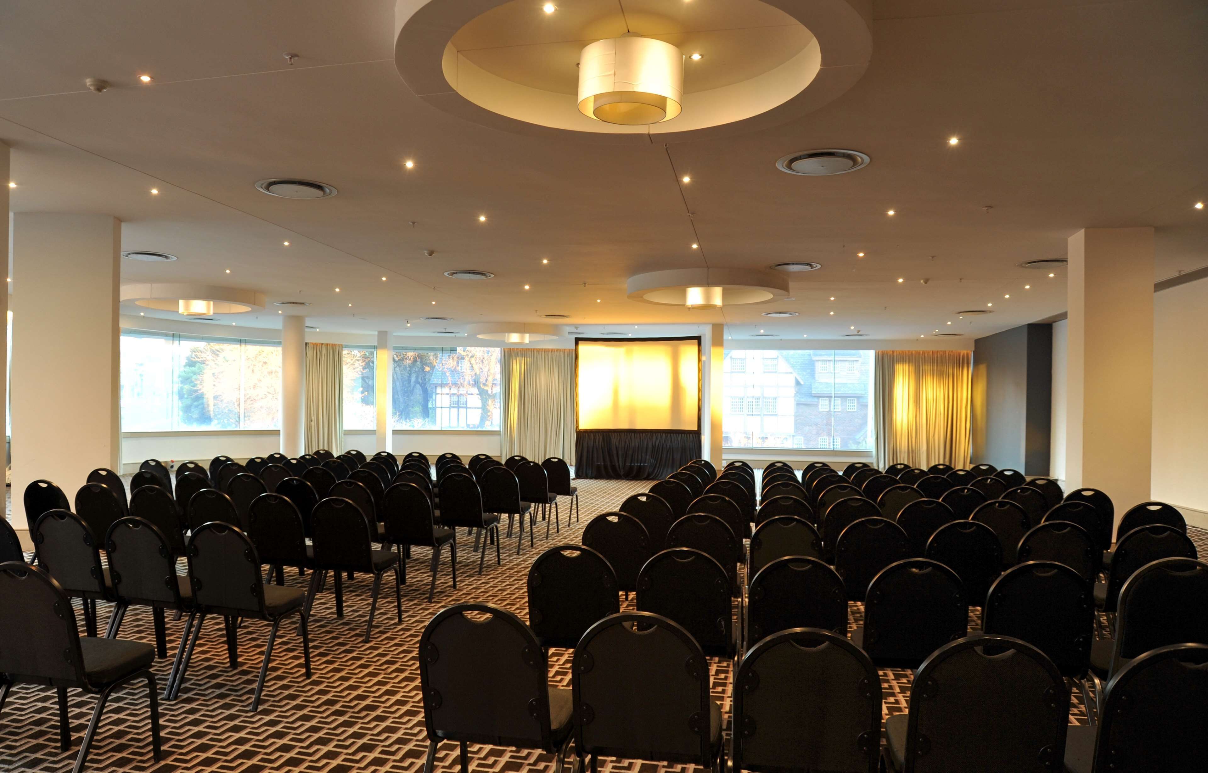 A conference room with rows of chairs, a stage, and large windows with curtains.
