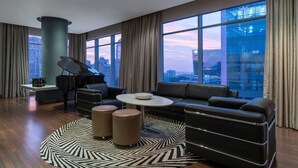 A modern living room with a grand piano, leather sofas, and a large window offering a city view.