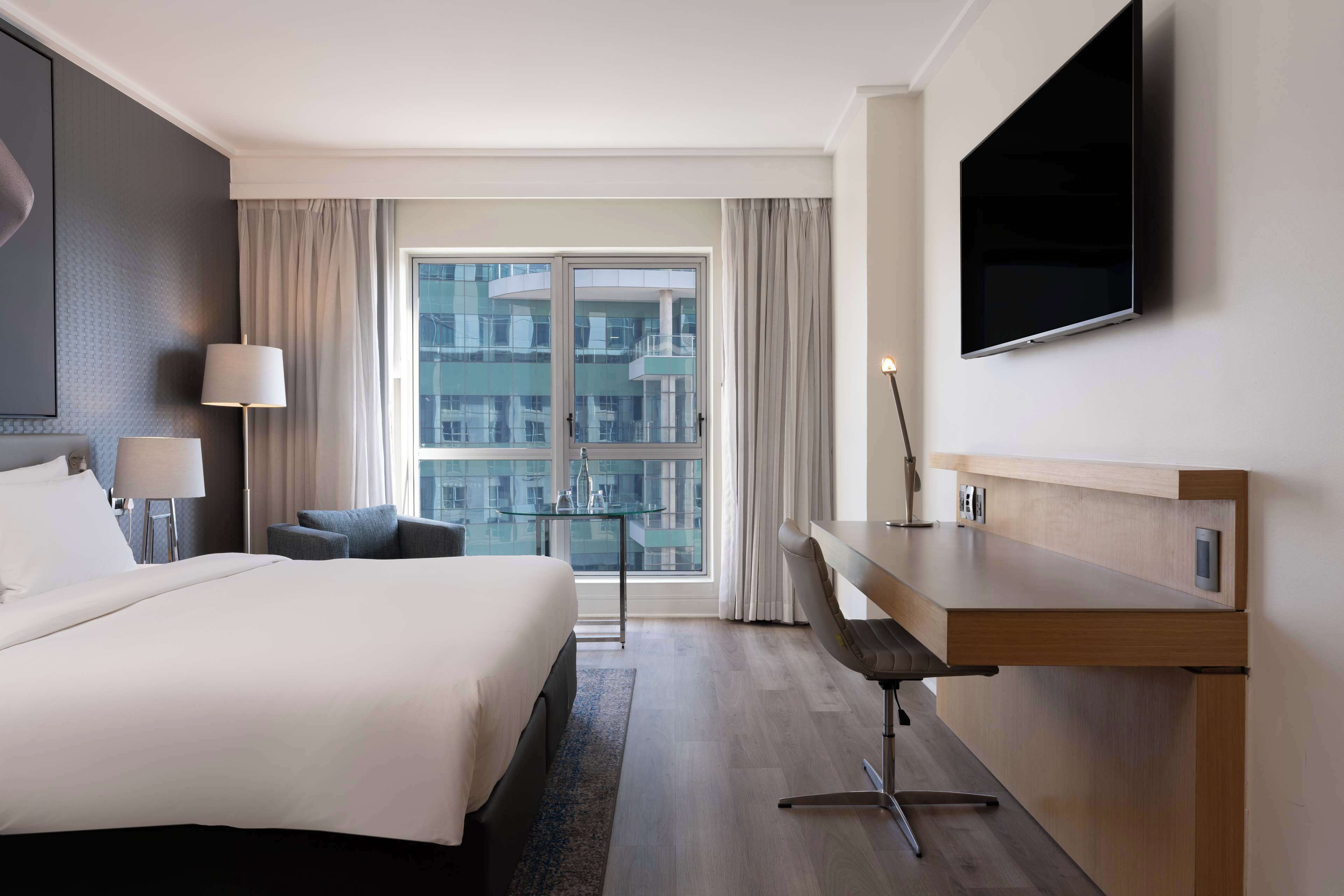 A modern hotel room with a large bed, a desk, a chair, a TV, and a window with curtains.
