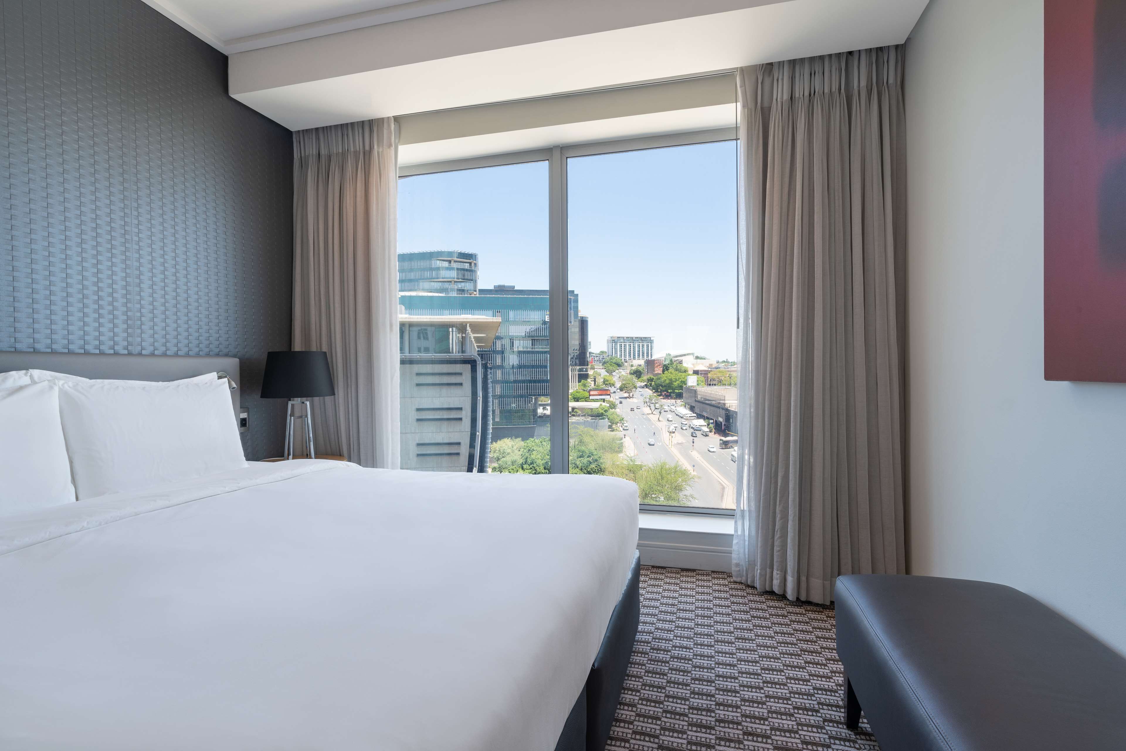 Une chambre d’hôtel avec un grand lit, un bureau, une chaise et une vue sur le paysage urbain grâce à de grandes fenêtres.