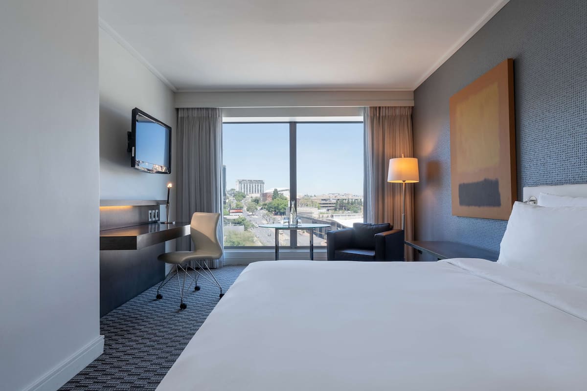 Une chambre d’hôtel avec un grand lit, un bureau avec une chaise, un téléviseur fixé au mur, une fenêtre donnant sur la ville et un tableau accroché au mur.