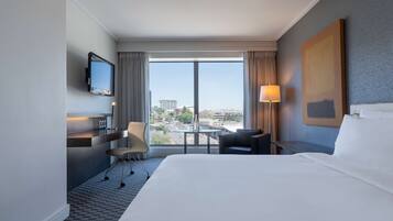 Quarto de hotel com uma cama grande, uma escrivaninha com cadeira, televisão na parede, janela com vista para a cidade e um quadro na parede.