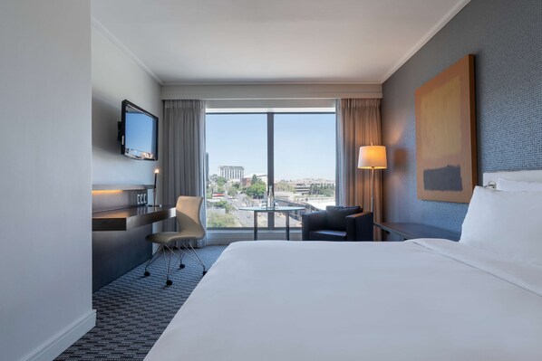 Habitación de hotel con una cama grande, un escritorio con silla, un televisor montado en la pared, una ventana con vista a la ciudad y un cuadro en la pared.