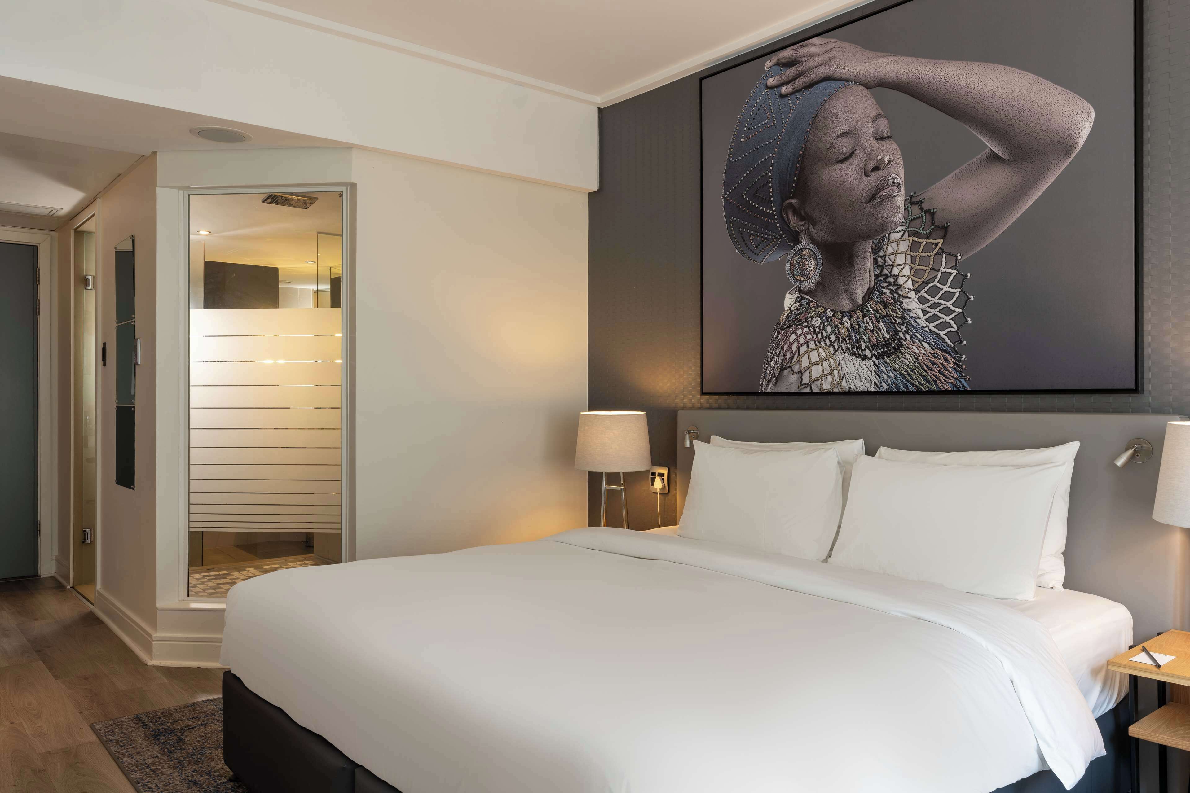 Una habitación de hotel con una cama grande, una mesita de noche con lámpara, un cuadro en la pared y una puerta que da a un pasillo.
