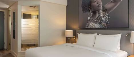 Una habitación de hotel con una cama grande, una mesita de noche con lámpara, un cuadro en la pared y una puerta que da a un pasillo.