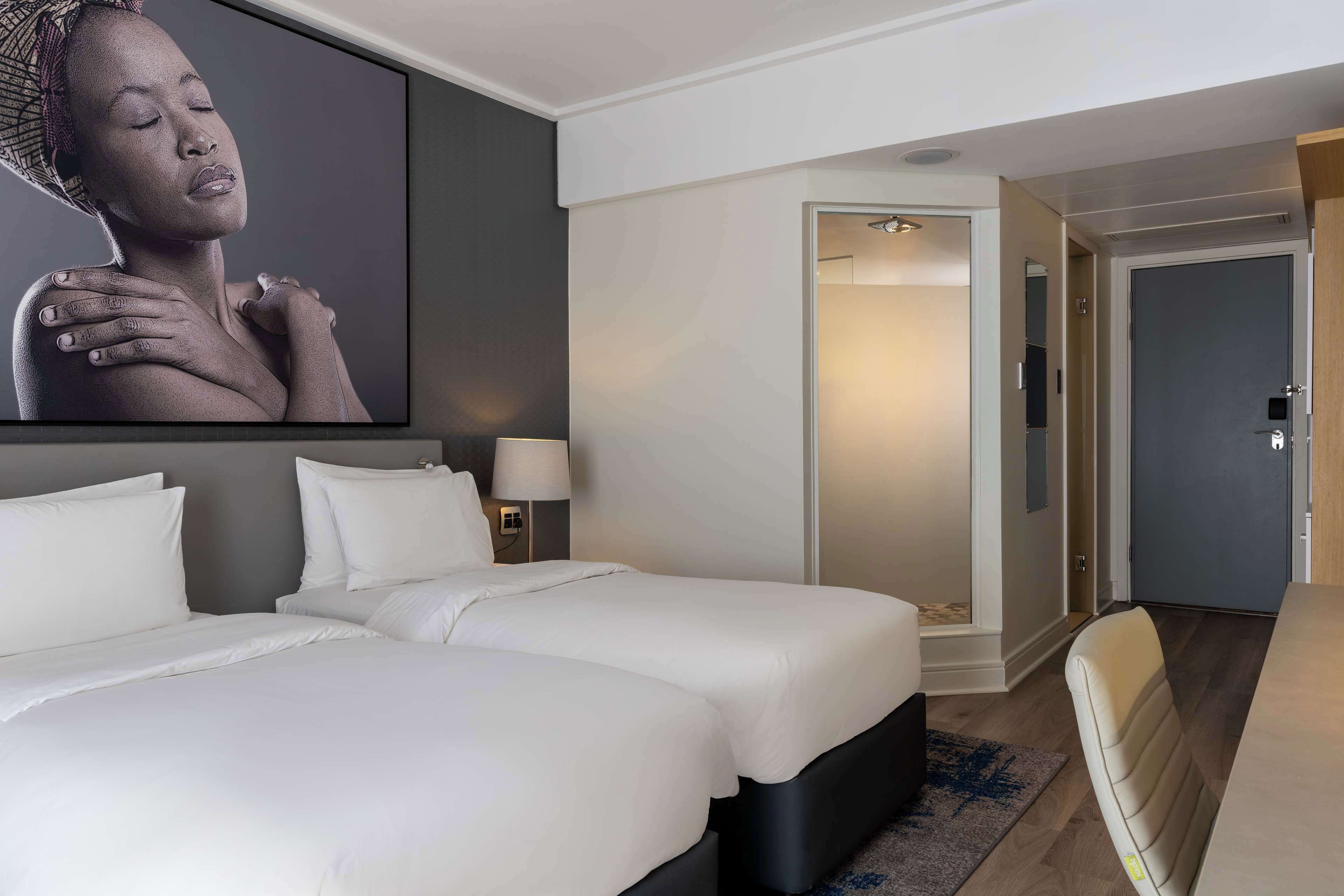 Habitación de hotel con dos camas, una obra de arte grande colgada en la pared, una mesita de noche con lámpara, y una puerta que da al baño.