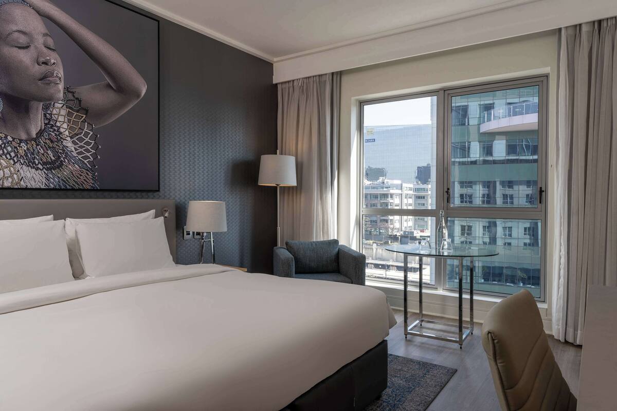 Une chambre d’hôtel avec un grand lit, une petite table, une chaise, une lampe et une grande fenêtre donnant sur la ville.