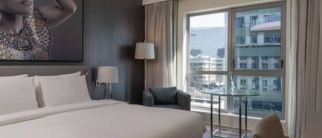A hotel room with a large bed, a small table, a chair, a lamp, and a large window with a city view.