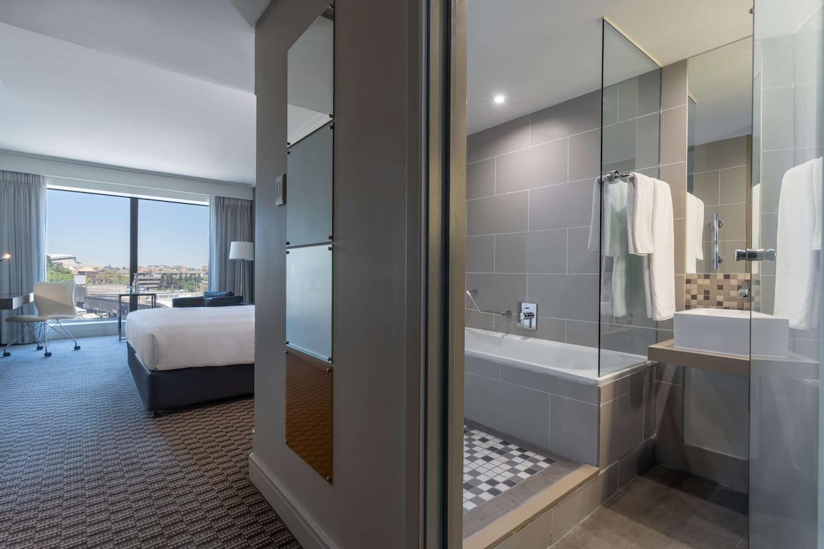 Une chambre d’hôtel moderne dotée d’un grand lit, d’une salle de bain séparée avec une douche à parois de verre, et offrant une vue sur la ville grâce à de grandes fenêtres.