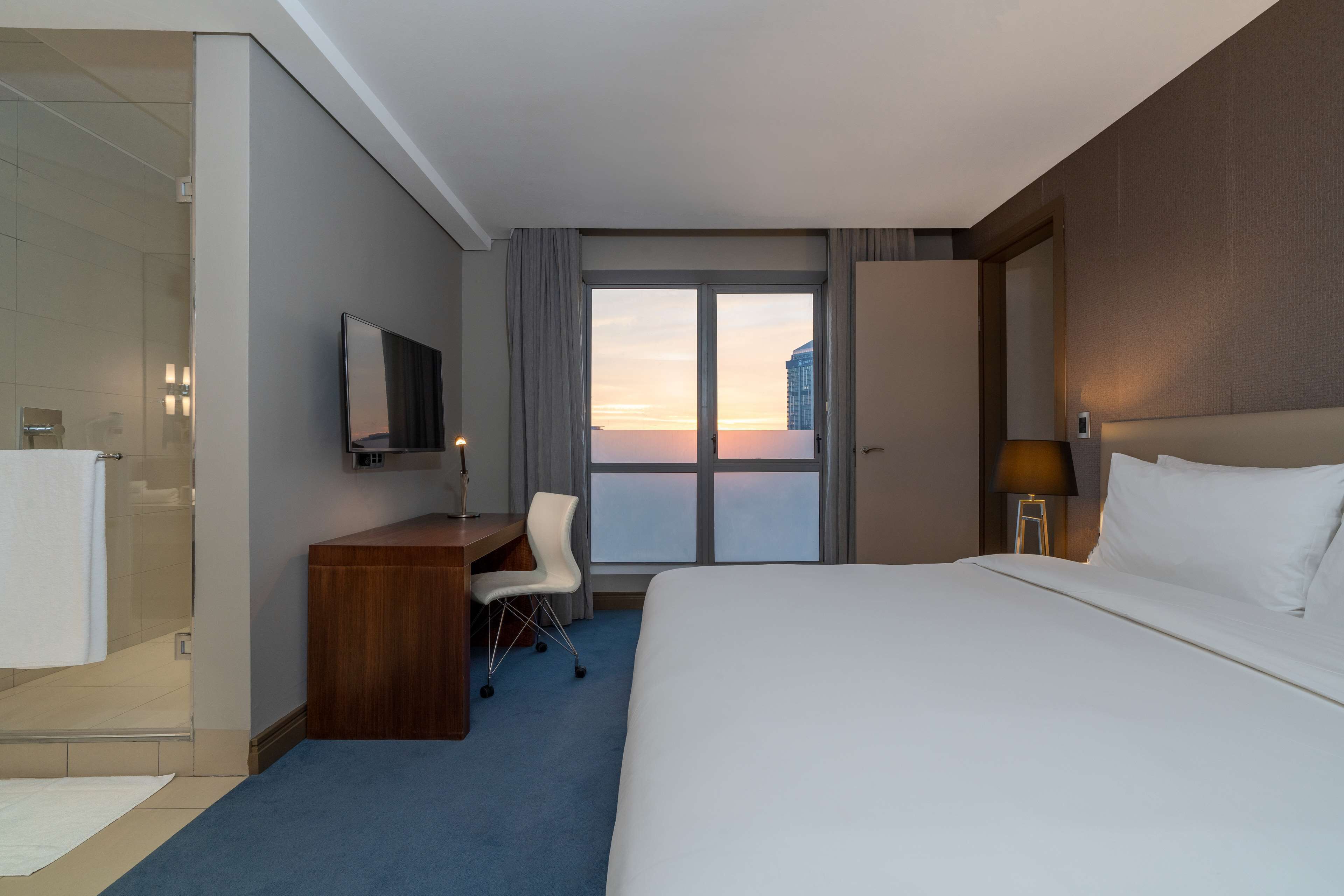 A hotel room with a bed, a desk, a chair, a TV, a window with a view, and a bathroom with a shower.