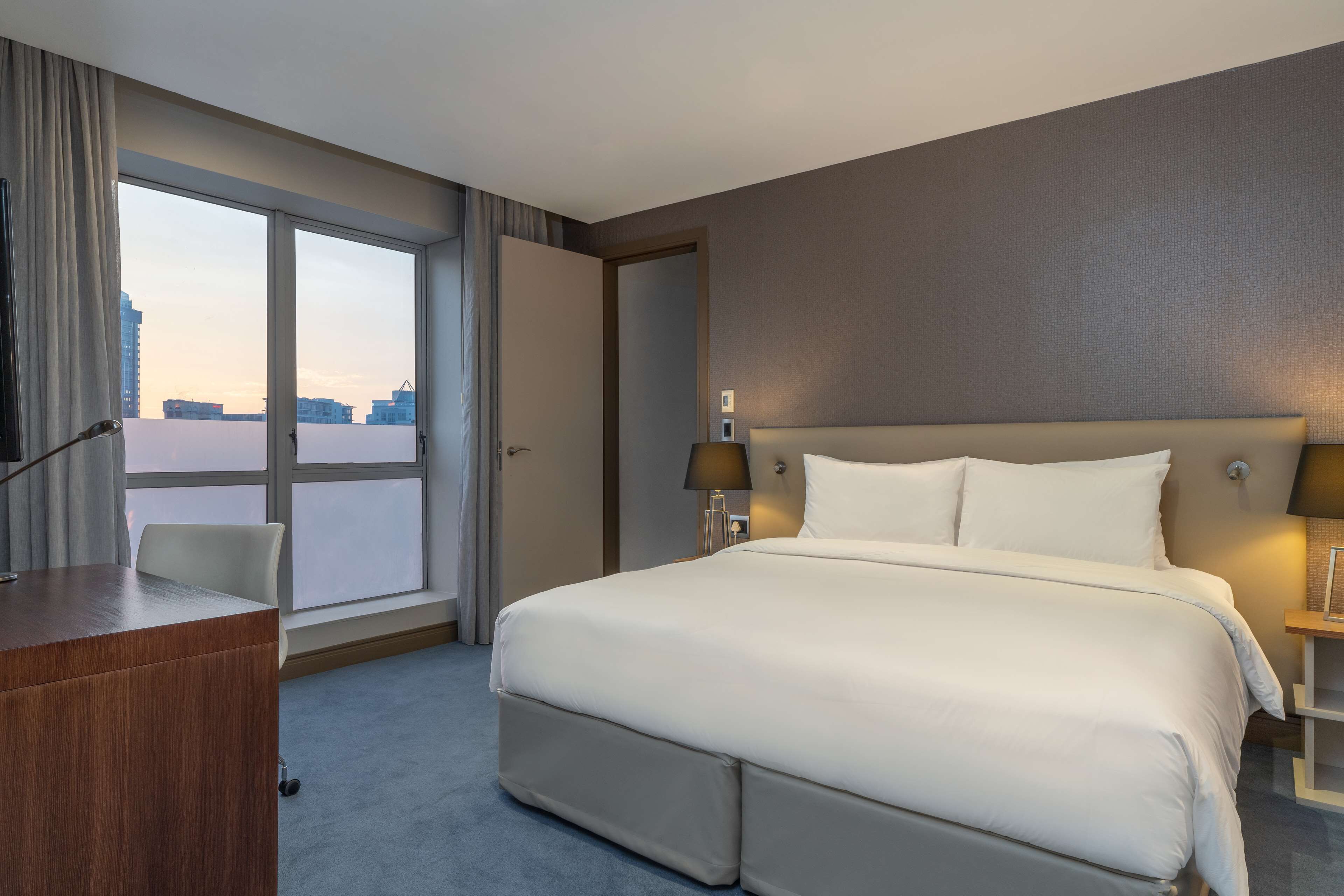 A hotel room with a large bed, a desk, a chair, and a view of the cityscape through the window.