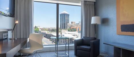A hotel room with a large window, a desk, a chair, a sofa, and a lamp.