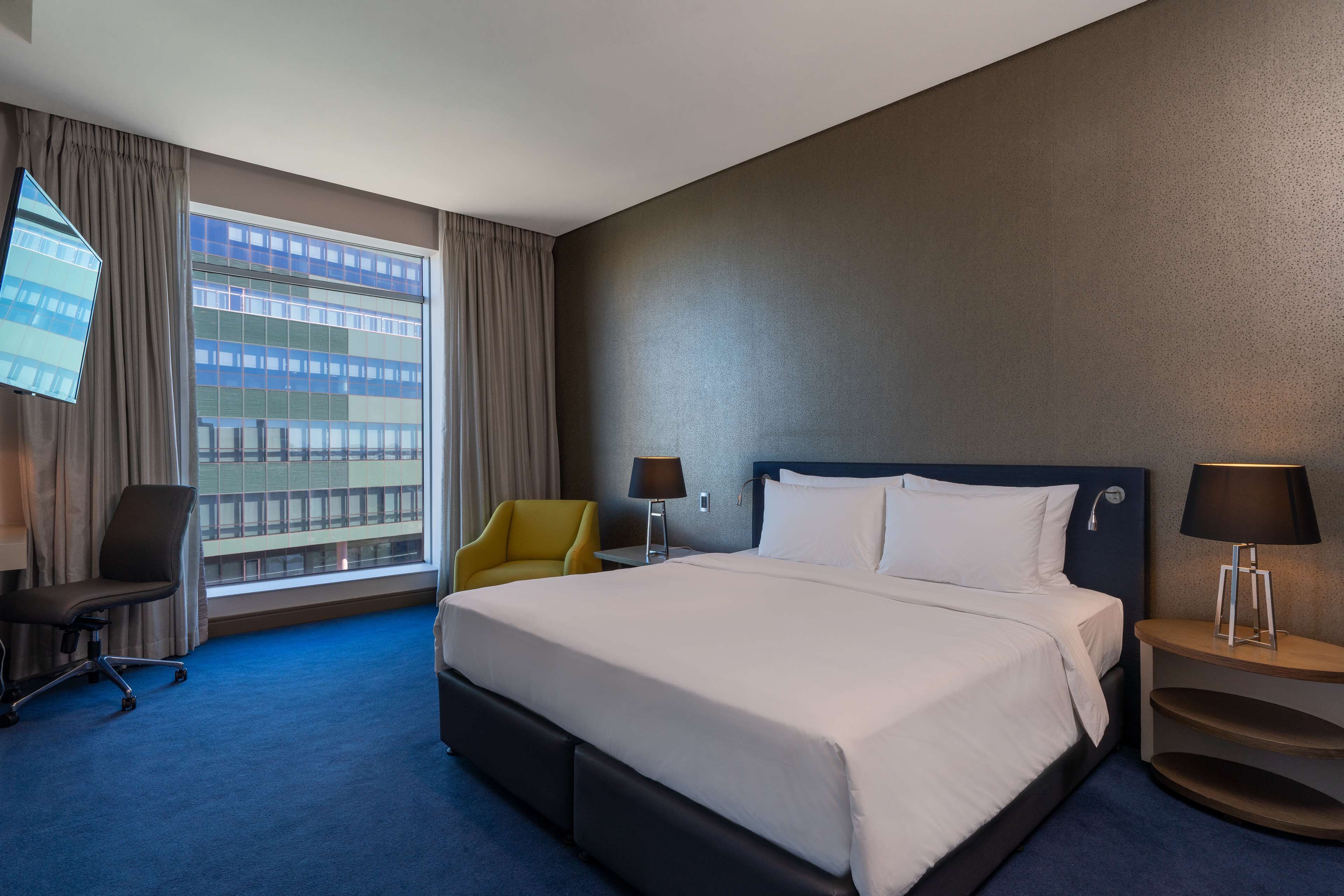 Habitación de hotel con una cama grande, un escritorio con lámpara, una silla y un televisor montado en la pared.