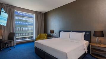 Habitación de hotel con una cama grande, un escritorio con lámpara, una silla y un televisor montado en la pared.