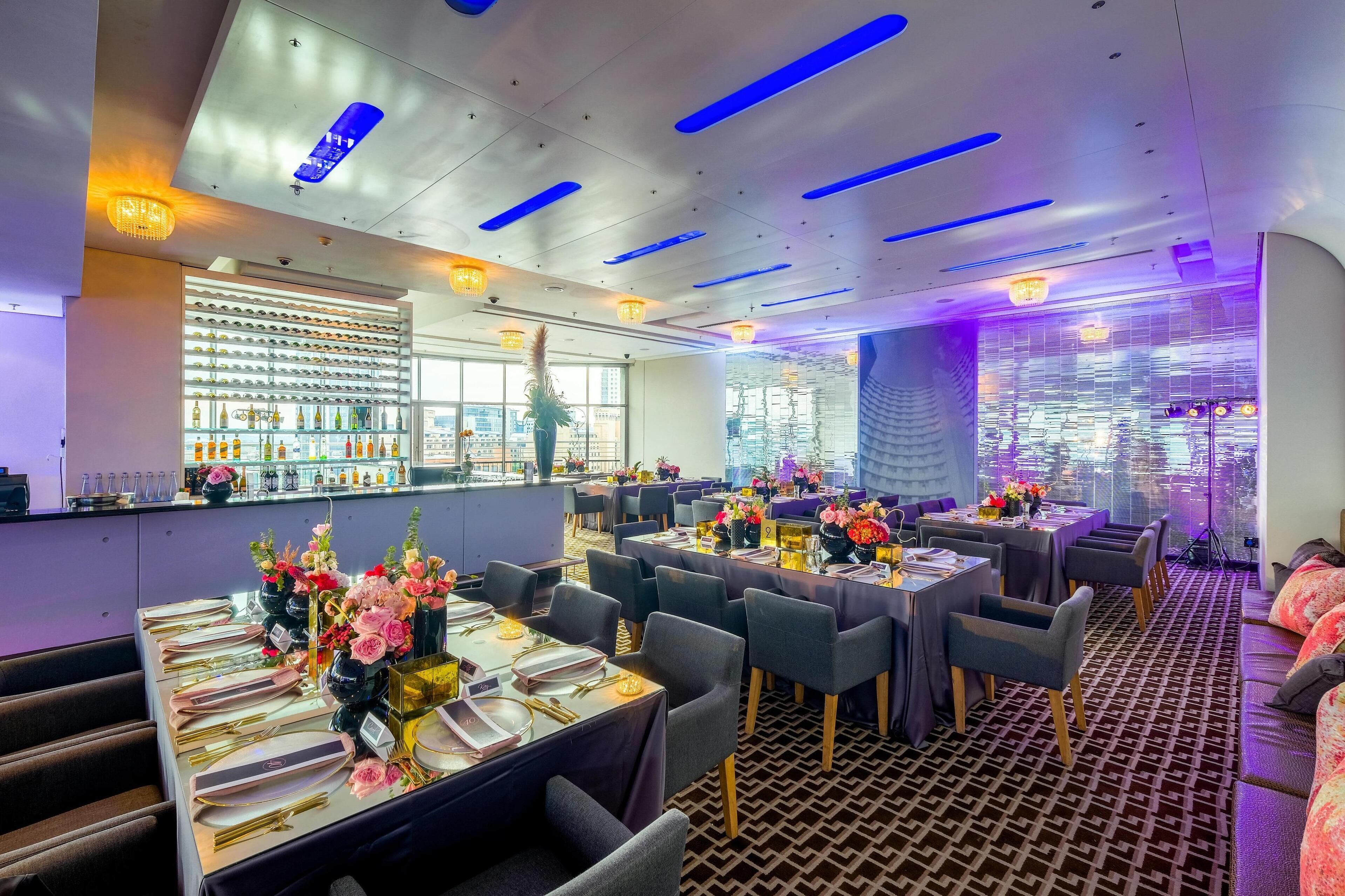 A modern restaurant with tables set for dining, a bar area with bottles, and a city view.