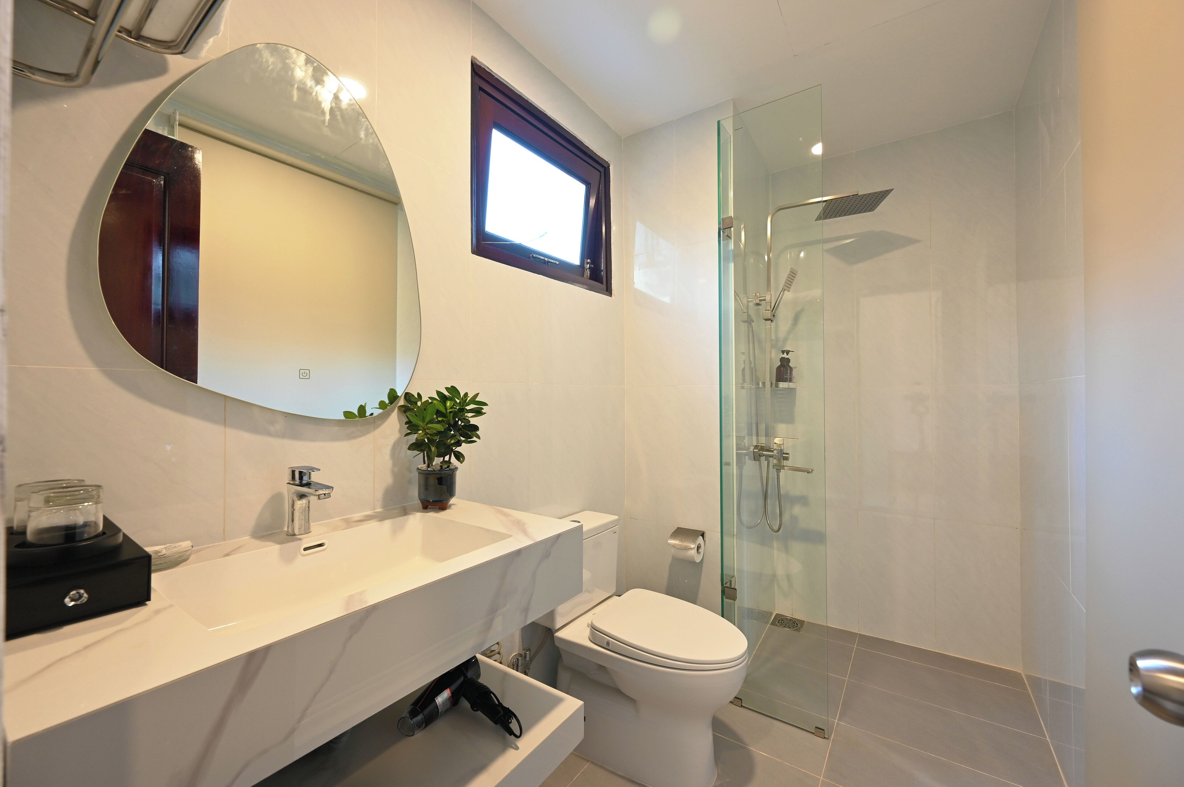Une salle de bain moderne avec un lavabo blanc, un miroir et des toilettes. On y trouve une douche en verre et une plante en pot sur le meuble-lavabo.