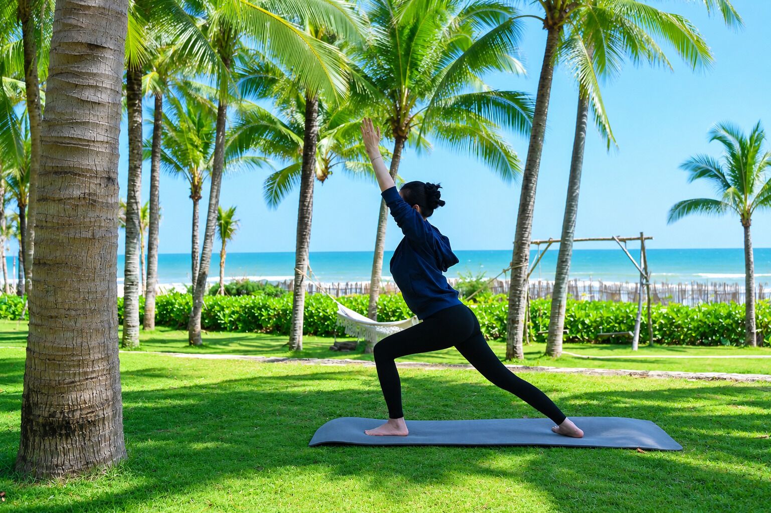 Una persona realizando una postura de yoga sobre una colchoneta en un área con césped, palmeras y vista al océano.