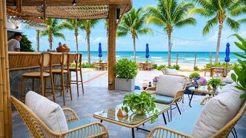 Un área de estar al aire libre frente a la playa, con mobiliario de mimbre, un bar y palmeras.
