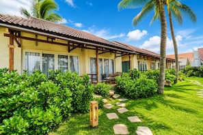 Un hotel con un área de estar cubierta al aire libre, un jardín bien cuidado con palmeras y un cielo despejado.