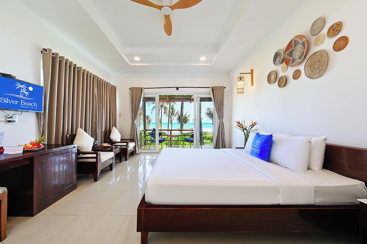 A hotel room with a bed, a desk, a TV, and a view of the beach.