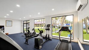 A gym with treadmills, elliptical machines, and stationary bikes, large windows with a view of palm trees and a building outside. - Silver Beach Resort Ke Ga (Tan Thanh)
