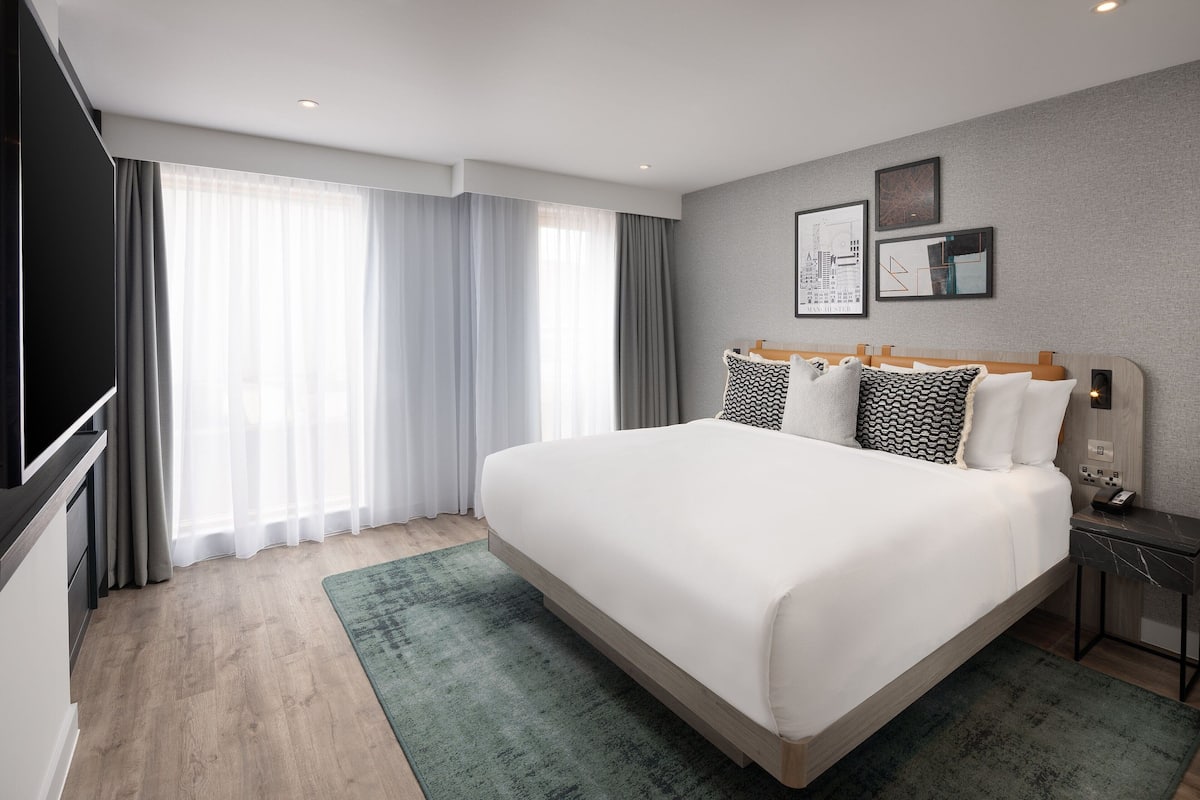 A modern hotel room with a large bed, a flat-screen TV, a bedside table, and a large window with curtains.