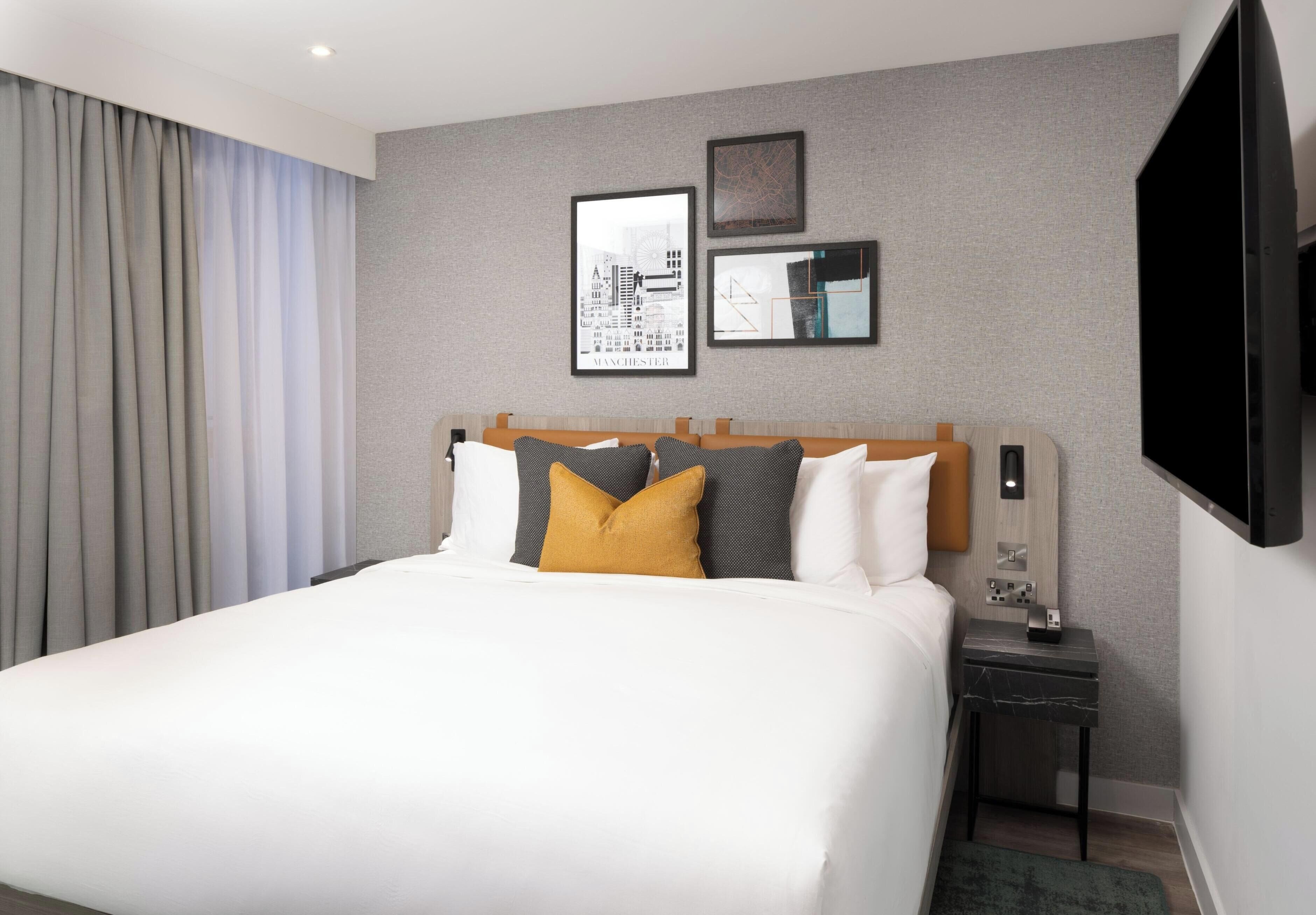 A hotel room with a large bed, a nightstand, a flat-screen TV, and a cityscape artwork on the wall.