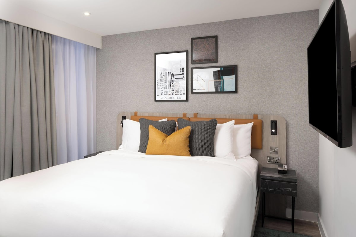 A hotel room with a large bed, a nightstand, a flat-screen TV, and a cityscape artwork on the wall.