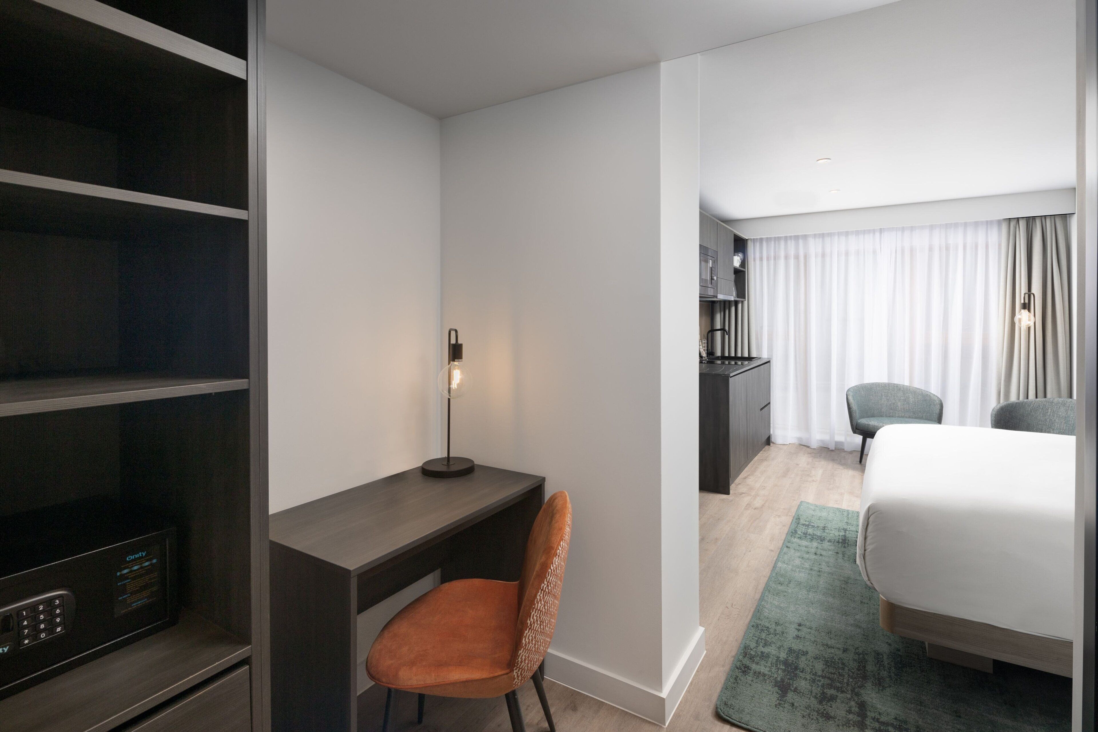 Quarto de hotel com escrivaninha, cadeira, cama e uma pequena cozinha equipada.