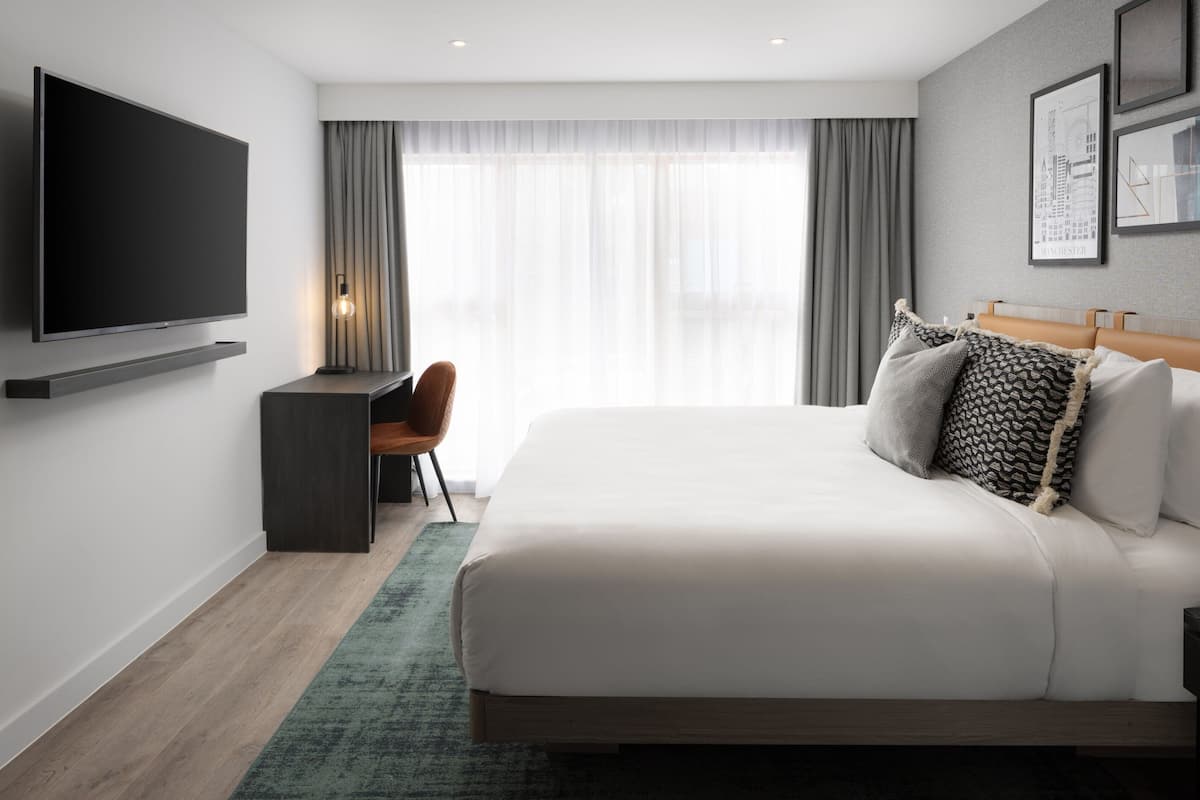 A modern hotel room with a large bed, a flat-screen TV mounted on the wall, a desk with a chair, and a view of the city through sheer curtains.