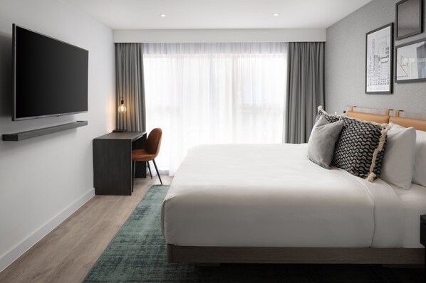 A modern hotel room with a large bed, a flat-screen TV mounted on the wall, a desk with a chair, and a view of the city through sheer curtains. - Residence Inn by Marriott Manchester Piccadilly (Manchester)