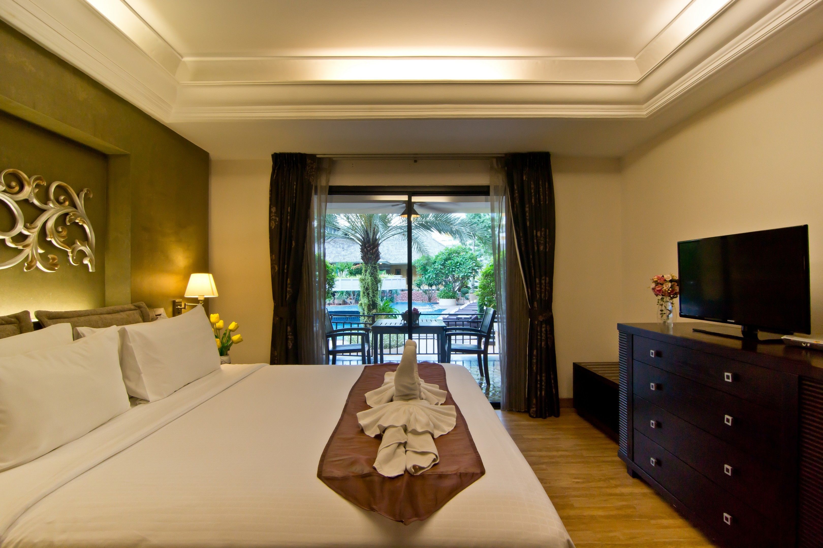 A hotel room with a neatly made bed, a television, a dresser, and a view of the outdoors through a glass door.