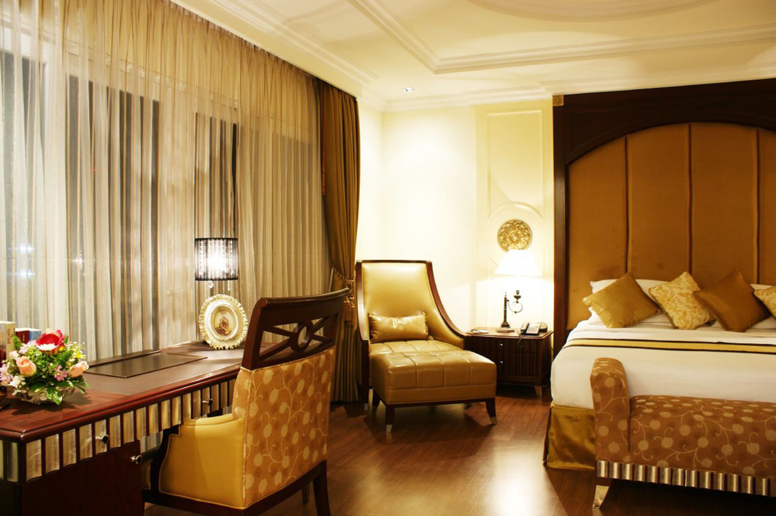 A hotel room with a large bed, a desk, two armchairs, and a nightstand with a lamp.