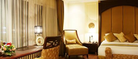 A hotel room with a large bed, a desk, two armchairs, and a nightstand with a lamp.