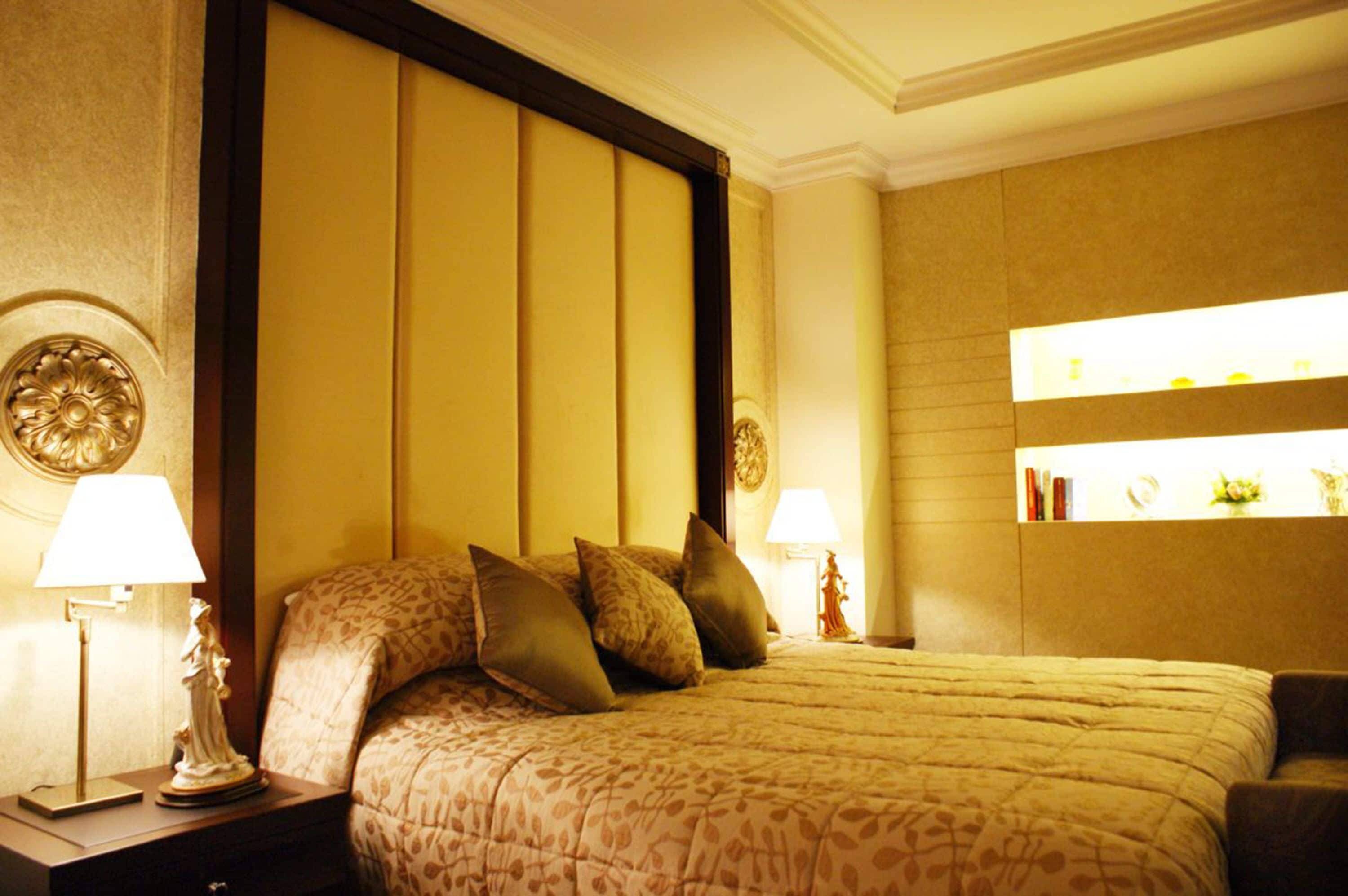 Une chambre d’hôtel avec un grand lit, deux tables de chevet avec des lampes, une étagère murale agrémentée d’objets décoratifs et une couvre-lit à motifs.