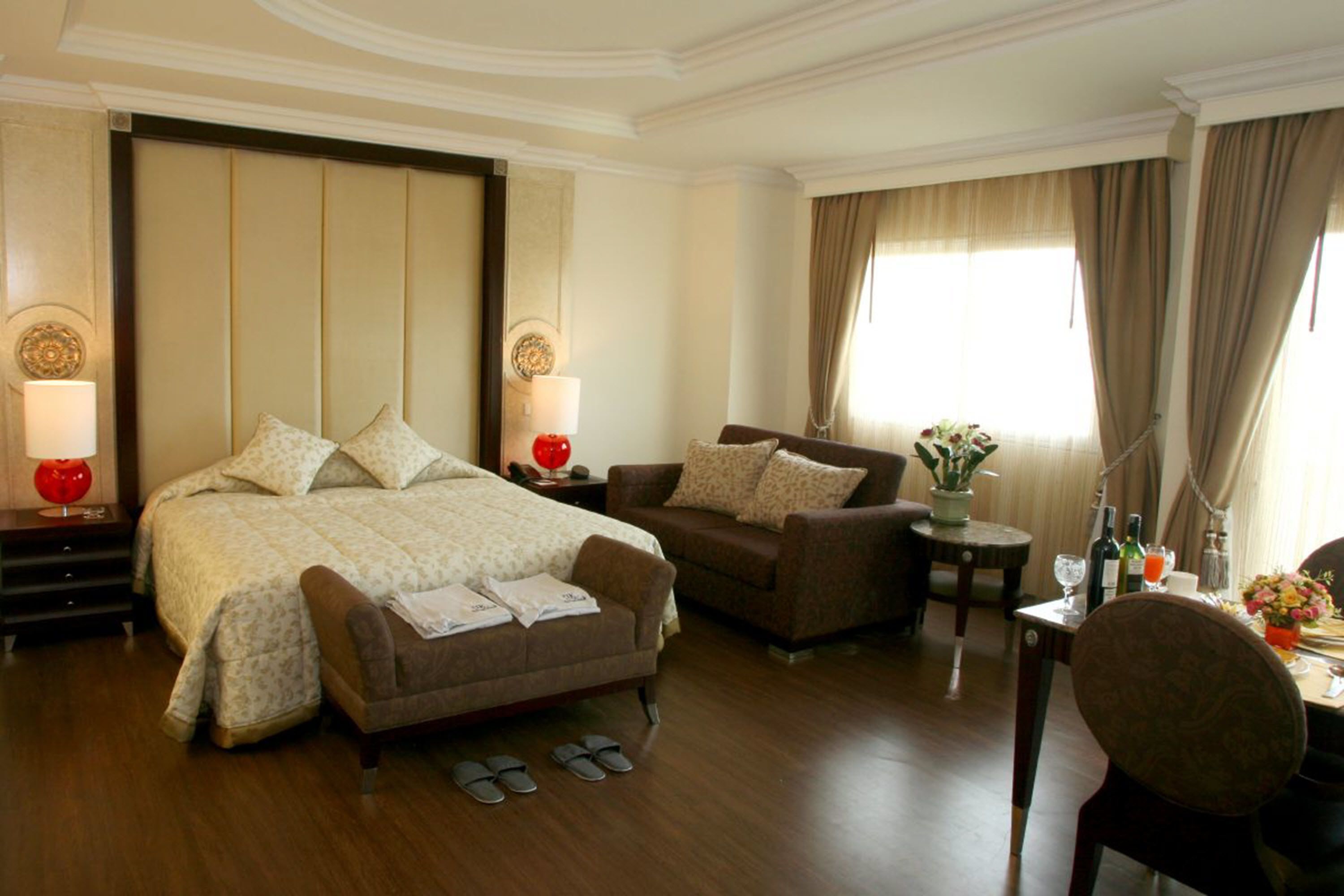 A hotel room with a large bed, two armchairs, a small table with a vase of flowers, and a desk with a lamp.