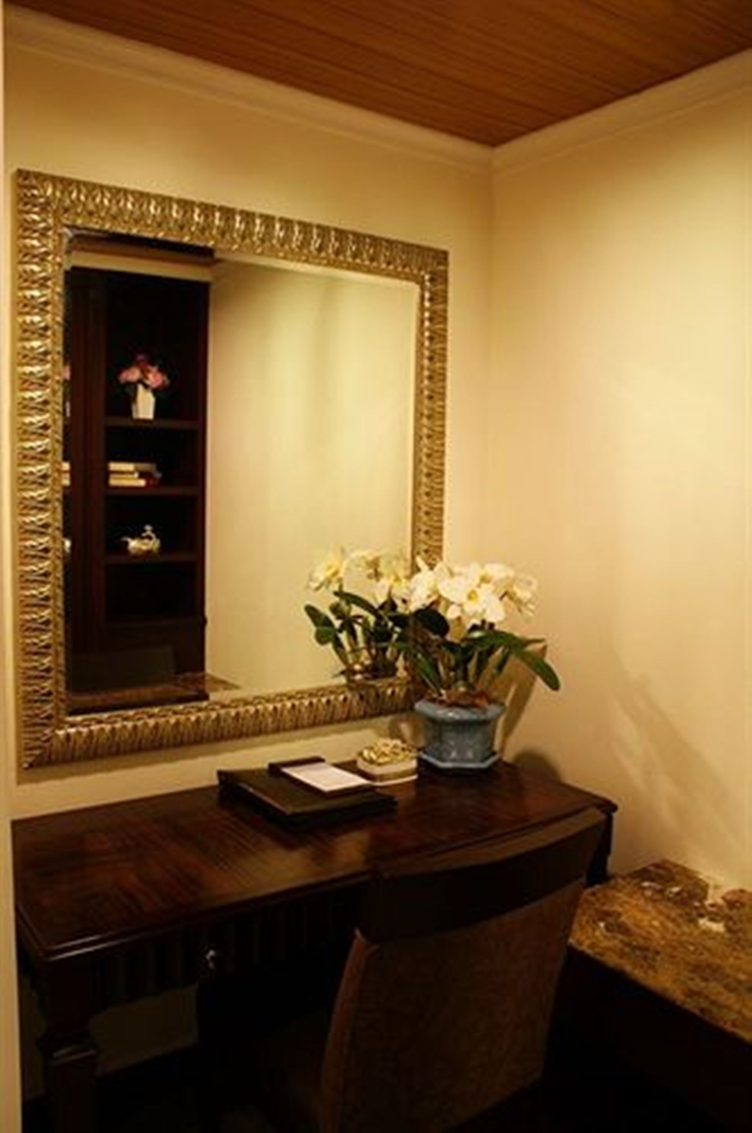 A desk with a mirror, a vase with flowers, and a shelf with books and a teapot.