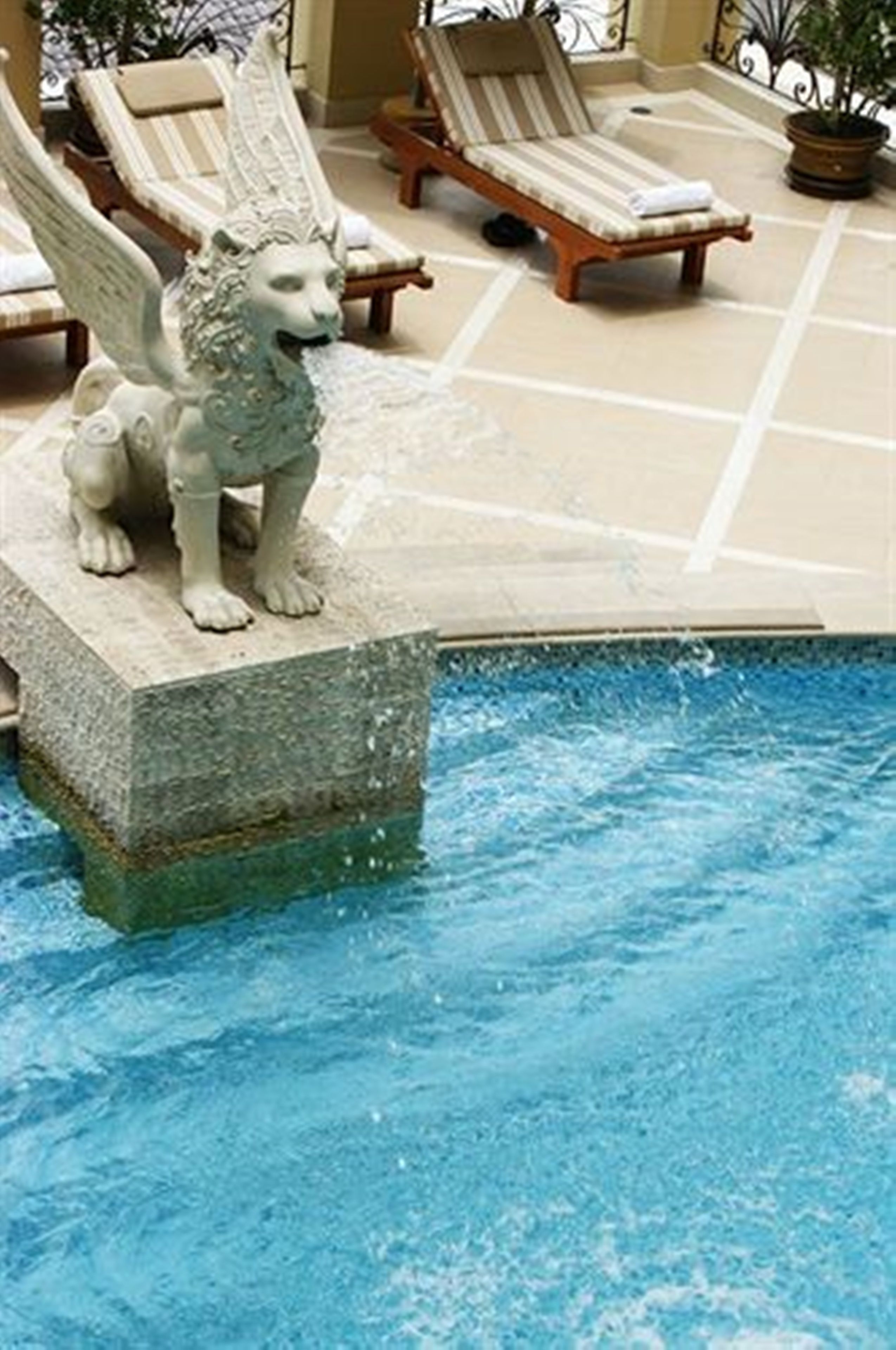 A stone statue of a winged lion by a poolside, lounge chairs, and potted plants.