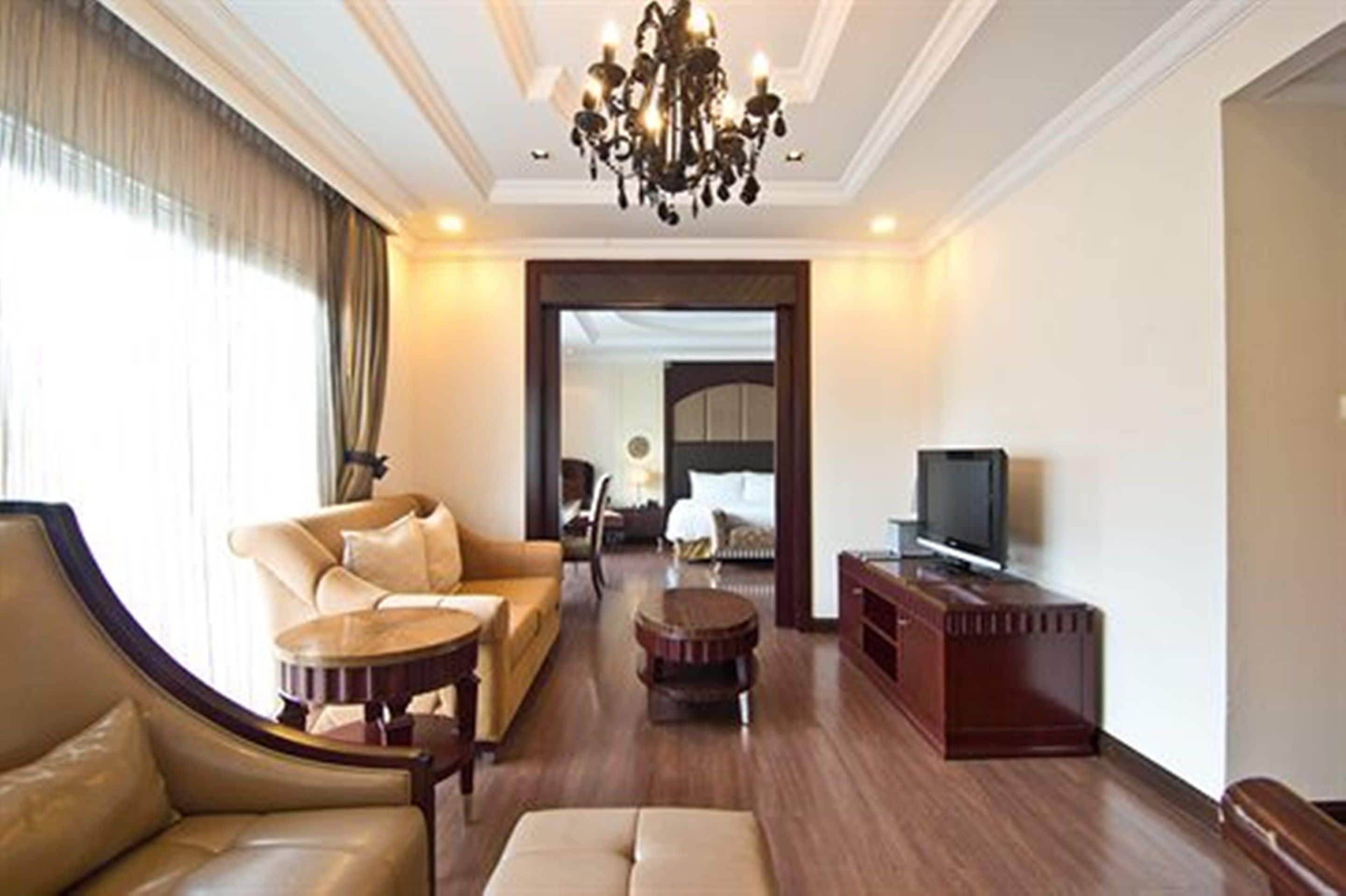 A spacious living room with a sofa, armchair, and ottoman, a television on a wooden stand, and a chandelier.