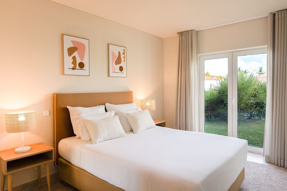 A hotel room with a large bed, two bedside lamps, and a view of greenery through the window.