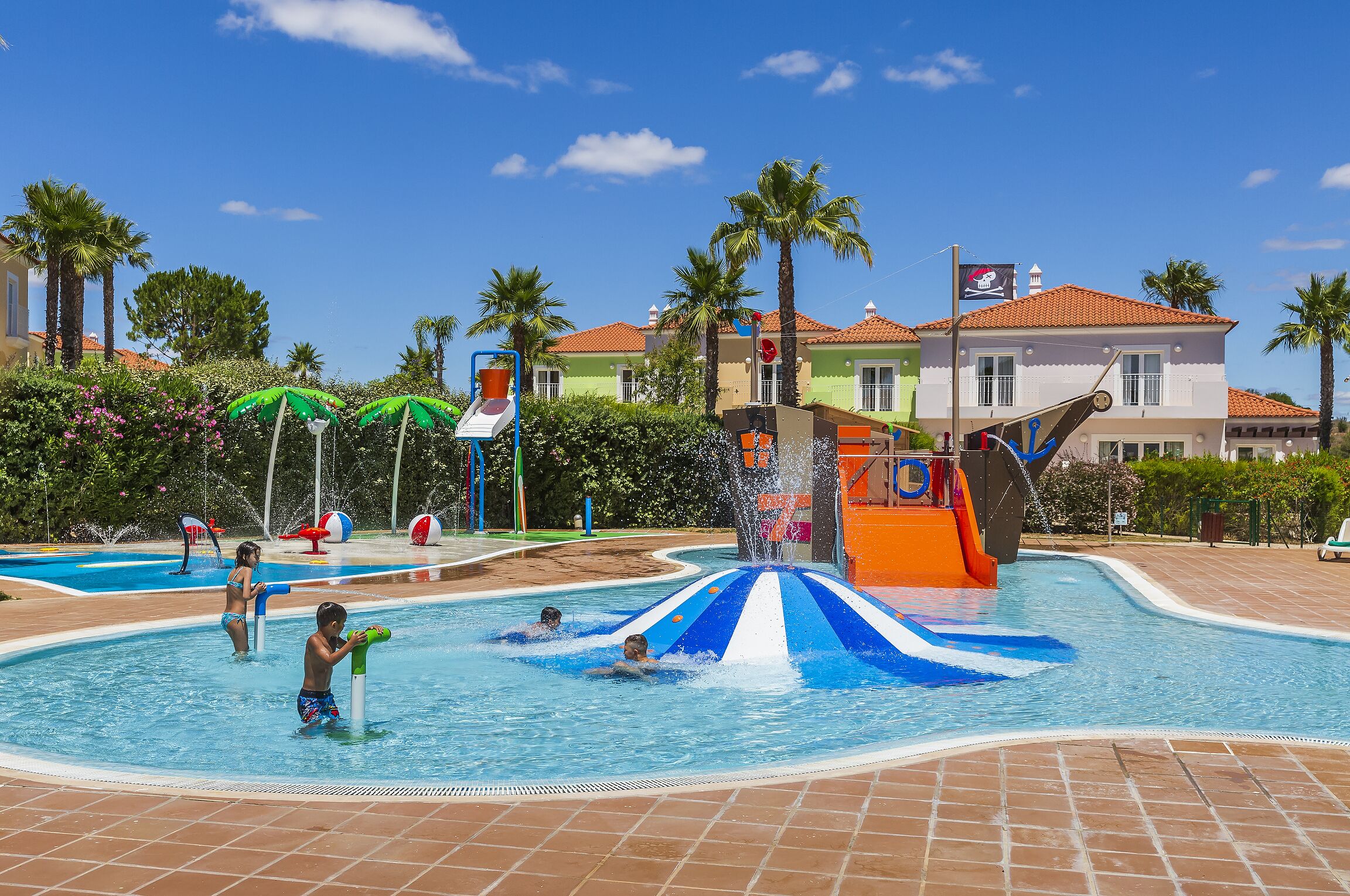 A swimming pool area with a water slide, palm trees, and a building with a red-tiled roof.