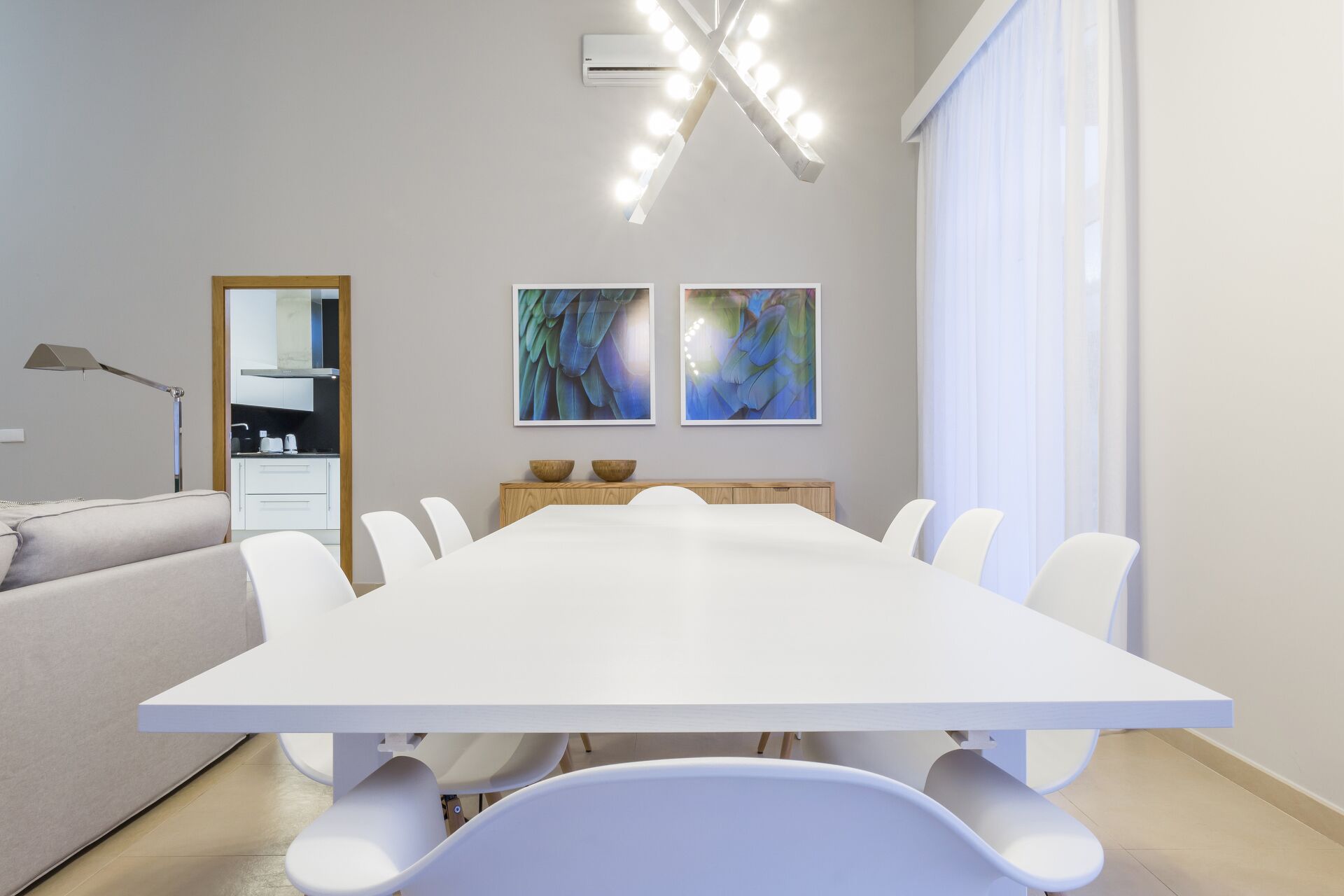 A modern dining room with a large white table, white chairs, and two abstract paintings on the wall.