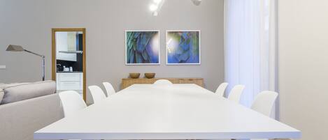 A modern dining room with a large white table, white chairs, and two abstract paintings on the wall.