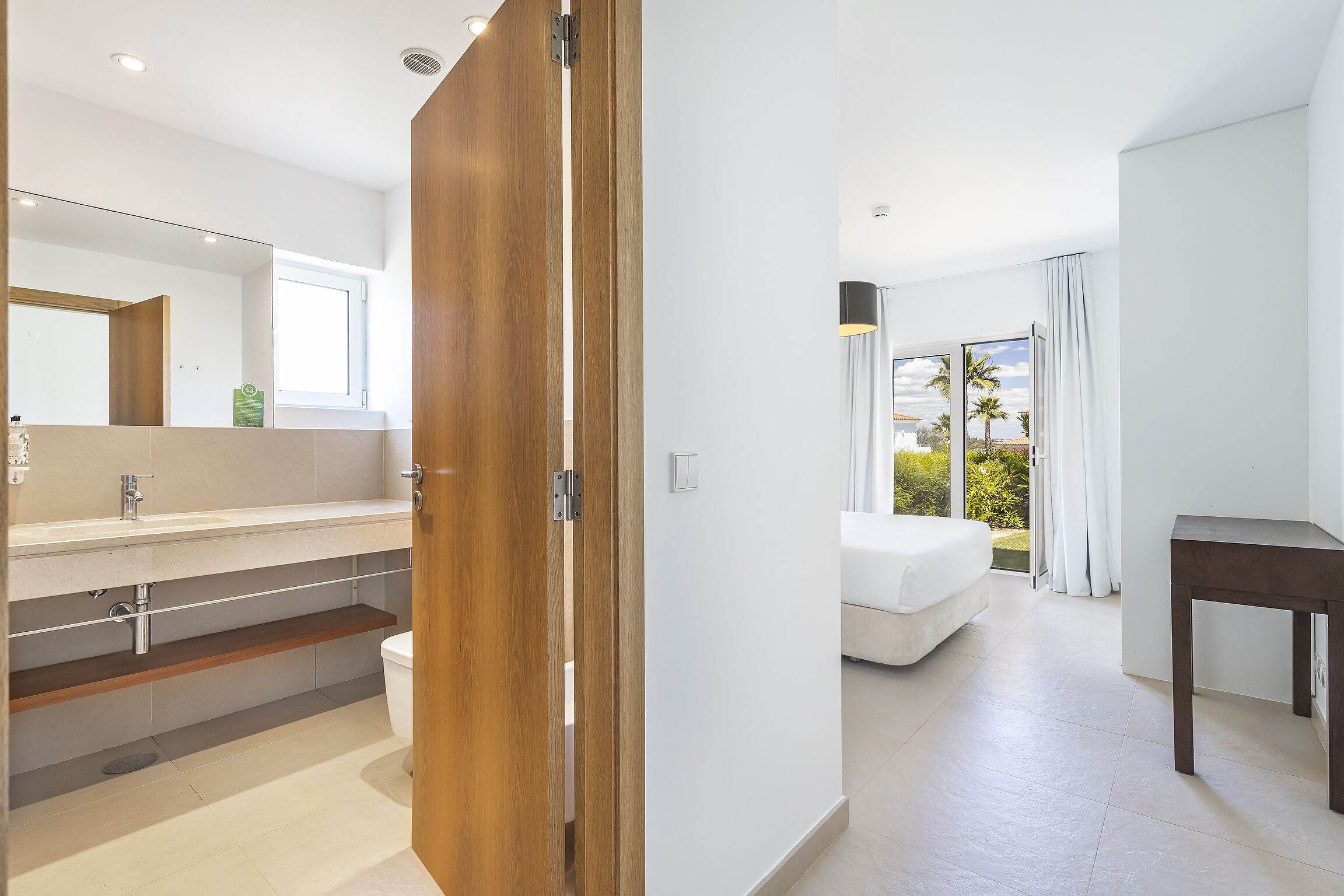 Moderni hotellihuone, jossa on puinen ovi, suuri peili, valkoinen sänky ja näkymä ulos.