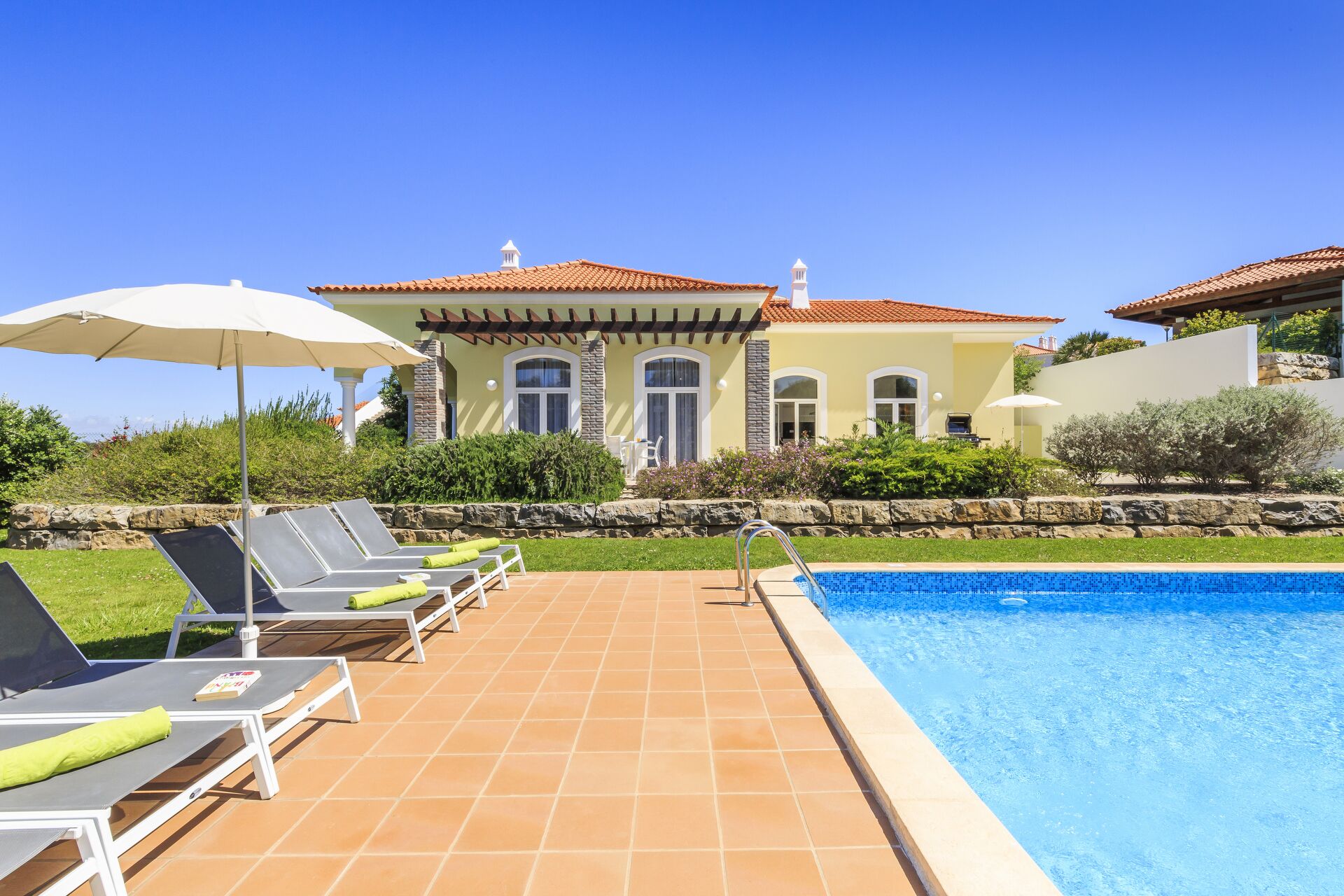 A poolside area with lounge chairs and umbrellas, a swimming pool, and a house with a tiled roof and stone walls.