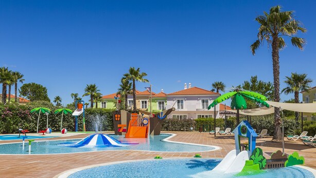 Zona de piscina con toboganes y fuentes, rodeada de palmeras y sillas de descanso.