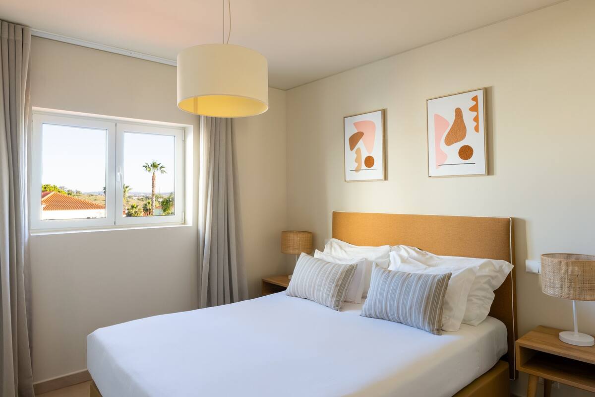 A hotel room with a large bed, two bedside lamps, a window with curtains, and two framed artworks on the wall.