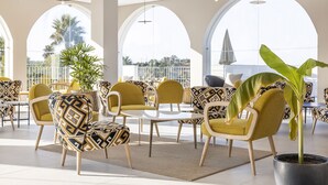 Un área de estar moderna en el interior, con sillas y mesas estampadas, ventanas grandes con marcos arqueados y una planta en maceta.